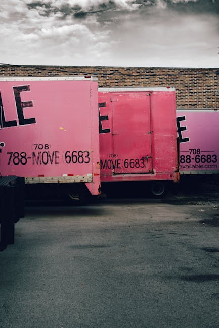 Image showing two pink moving containers parked against a brick wall during daytime with cloudy skies. The containers are made of metal and are positioned on a paved surface, with one slightly overlap