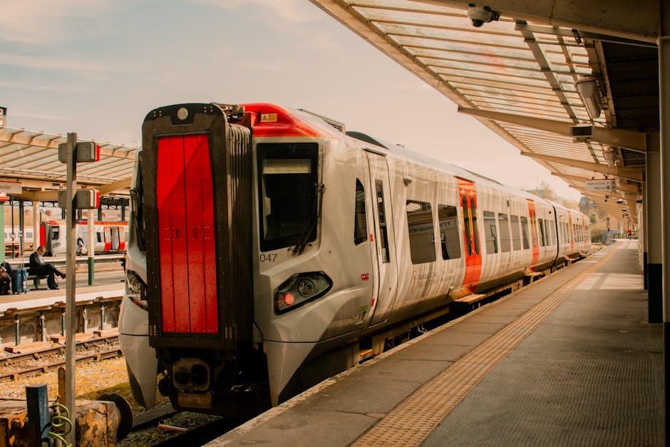 A modern, silver and red train is stationary at East Croydon Station platform during daylight, with the platform roof overhead featuring a metal and glass canopy. The train's front section and side windows are visible, along with external safety markings. In the background, there are other trains and passengers waiting on the adjacent platform, some sitting on benches or walking. Luggage and moving boxes, wrapped in cardboard and plastic, are seen on the platform, suggesting preparations for home relocation or furniture transport. The scene captures the loading process typical of moving services provided by Removal Companies Croydon, with the train ready for passenger boarding or cargo transfer as part of smooth moving logistics connected to house removals or furniture transport in the Croydon area.
