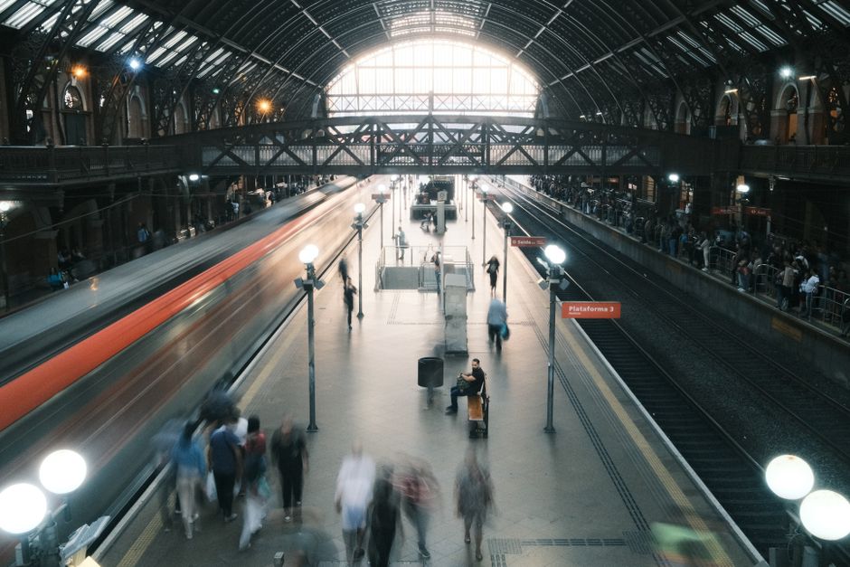 Inside East Croydon Station, the image shows a busy platform with commuters walking and waiting for trains. The station features a large arched glass ceiling allowing natural light to illuminate the area, which has a spacious design with multiple tracks and platforms. On the left side, a train is arriving or departing, captured with motion blur to indicate movement, while other passengers are seen standing or sitting on benches along the platform. There are street lamps providing additional lighting, and the station's architecture includes metal railings, brick walls, and elevated walkways visible in the background. Luggage, moving boxes, and wrapped furniture items are positioned near the station entrances, hinting at recent or upcoming home relocations. Moving equipment such as trolleys and straps may be seen assisting with the transport of household goods, consistent with professional removals services offered by Removal Companies Croydon, catering to those needing efficient furniture transport and packing during house moves.