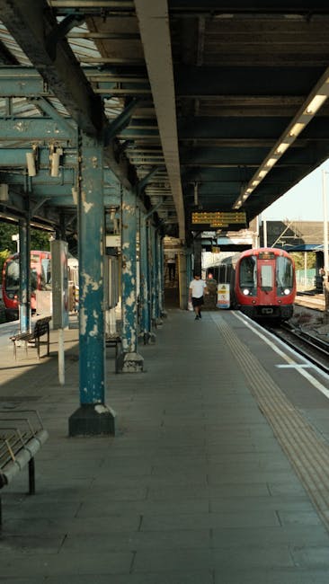 A modern, silver and red train is stationary at East Croydon Station platform during daylight, with the platform roof overhead featuring a metal and glass canopy. The train's front section and side windows are visible, along with external safety markings. In the background, there are other trains and passengers waiting on the adjacent platform, some sitting on benches or walking. Luggage and moving boxes, wrapped in cardboard and plastic, are seen on the platform, suggesting preparations for home relocation or furniture transport. The scene captures the loading process typical of moving services provided by Removal Companies Croydon, with the train ready for passenger boarding or cargo transfer as part of smooth moving logistics connected to house removals or furniture transport in the Croydon area.