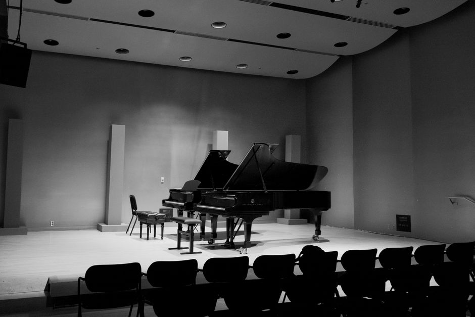 Interior view of a concert hall stage with a closed black grand piano positioned centrally, accompanied by three chairs and a small table, all surrounded by a wooden floor. The stage is bordered by a row of seats in the foreground and features minimalist acoustic panels on the wall behind the piano. The lighting is dim, highlighting the piano and seating area, with the stage prepared likely for a musical performance or rehearsal. This setting exemplifies a typical environment where professional removals companies, such as Removal Companies Croydon, might assist with packing, transporting, or moving musical instruments during a home relocation or event setup. The spacious and organized environment indicates careful handling of delicate items like pianos, suitable for the company's house removals and furniture transport services.