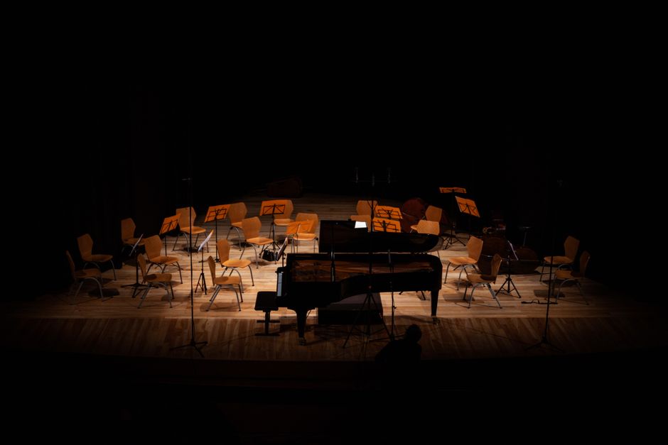 An empty stage at Fairfield Halls, Croydon, with a grand piano positioned centrally and surrounded by multiple light-colored wooden chairs arranged in a semi-circle, each with metal legs and some with sheet music stands. The stage is lit with focused lighting, creating a contrast against the dark background and black curtains. Wooden flooring is visible beneath the chairs and piano. This scene depicts a setup for a musical performance, possibly involving furniture and equipment arranged for a home or venue relocation, with the piano and chairs awaiting transport or reassembly as part of a professional removals process conducted by Removal Companies Croydon.