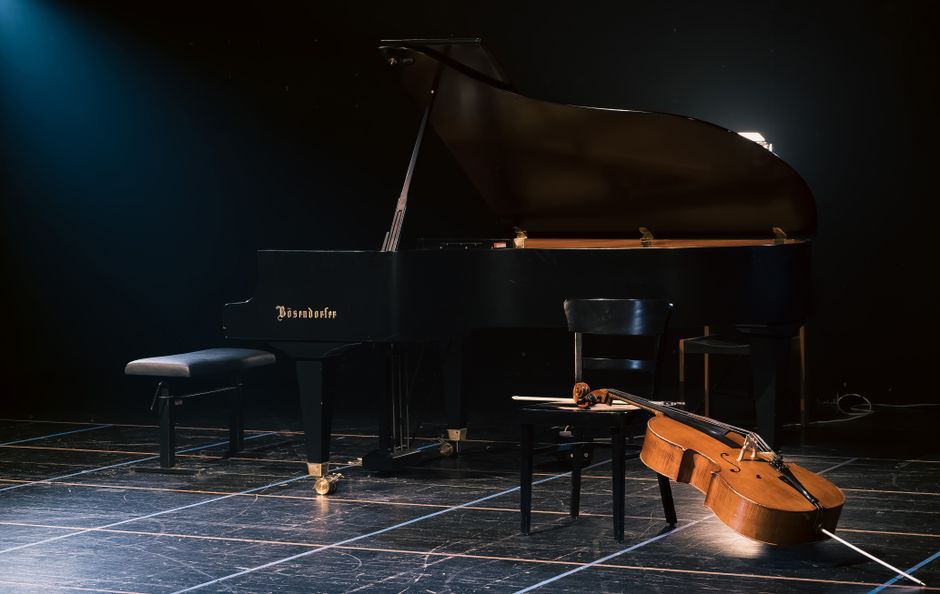 Interior view of a concert hall stage with a closed black grand piano positioned centrally, accompanied by three chairs and a small table, all surrounded by a wooden floor. The stage is bordered by a row of seats in the foreground and features minimalist acoustic panels on the wall behind the piano. The lighting is dim, highlighting the piano and seating area, with the stage prepared likely for a musical performance or rehearsal. This setting exemplifies a typical environment where professional removals companies, such as Removal Companies Croydon, might assist with packing, transporting, or moving musical instruments during a home relocation or event setup. The spacious and organized environment indicates careful handling of delicate items like pianos, suitable for the company's house removals and furniture transport services.