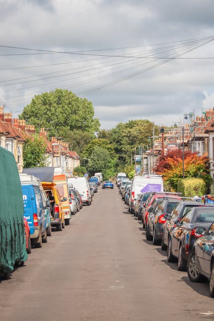 A residential street scene showing parked cars lining both sides of the road, with a variety of vehicles including small hatchbacks, vans, and larger delivery trucks. The street is paved with tarmac and features row houses with brick facades, tiled roofs, and small front gardens, some with trees and shrubs. Overhead, utility power lines stretch across the sky, which is overcast with grey clouds. In the background, the street continues with more parked vehicles and houses visible under natural daylight. The scene reflects a typical urban or suburban neighbourhood where home relocation or furniture transport may require careful planning to navigate tight drives. This setting illustrates the contextual environment where a professional removals service, such as [COMPANY_NAME], might operate during house removals or moving operations, often involving loading and unloading of furniture and boxes onto vans or trucks parked along the street.