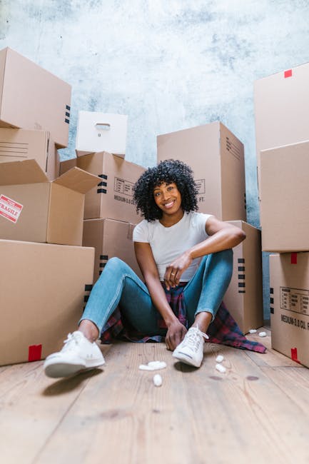 A young woman with curly black hair and wearing casual clothing, including a light grey t-shirt, blue jeans, and white sneakers, is sitting on a wooden floor in a room filled with stacked cardboard mo